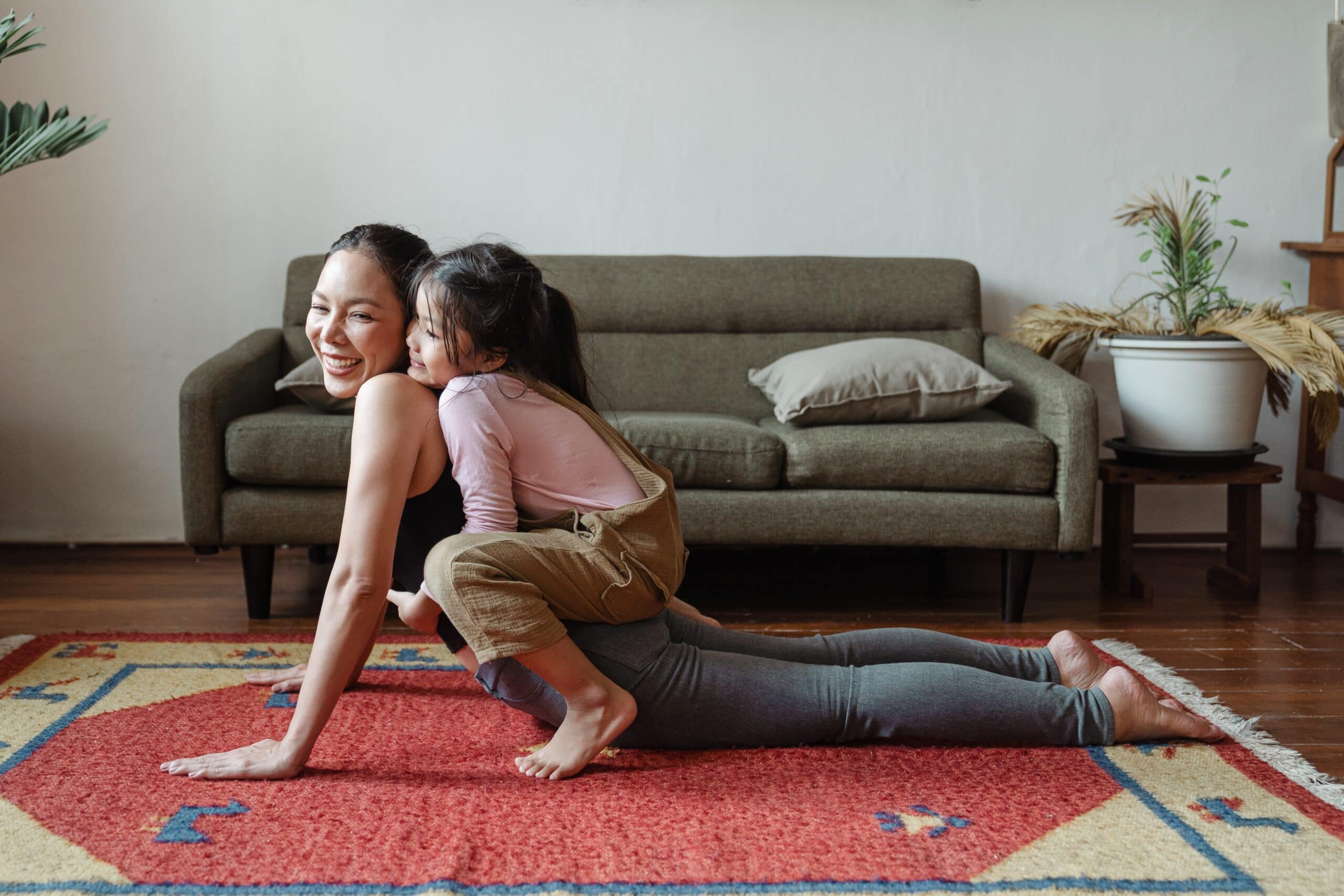 woman doing yoga stretch in her living room with her child hugging her back
