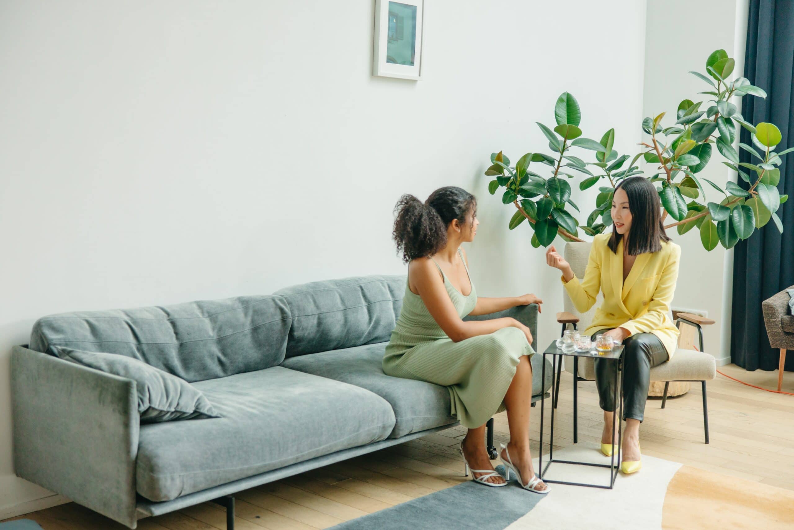 Woman in yellow blazer sitting in chair while talking to woman in sage green dress who is sitting on a couch